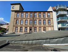 Backstein-Fassade eines dreistöckigen historischen Gebäudes mit mehreren Fenstern und einem kleinen Turm auf der linken Seite, daneben ein moderner Wohnblock mit blauen Balkonen. Im Vordergrund ein flaches, graues Dach.