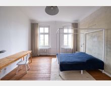 Helles Schlafzimmer mit Holzboden, einem Himmelbett mit dunkelblauer Decke, zwei großen Fenstern mit beigefarbenen Vorhängen und einem einfachen Holztisch mit weißem Stuhl. Eine Wand ist unverputzt, während die anderen weiß gestrichen sind.