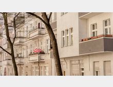 Fassaden mehrerer Wohngebäude in einer städtischen Straße, mit hellen Fassaden, Balkonen und einem Baum im Vordergrund. Ein Balkon zeigt einen rot-weiß gestreiften Sonnenschirm.