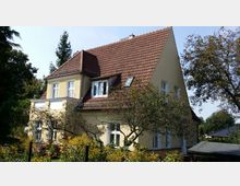 Zweigeschossiges Wohnhaus mit heller Fassade und einem steilen, roten Satteldach, umgeben von Gartenbäumen und blühender Vegetation. Im Vordergrund befindet sich ein Zaun, der das Grundstück begrenzt.