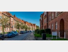 Straße mit zweistöckigen Wohnhäusern aus rotem Backstein, großen Fenstern und Giebeldächern. Entlang der gepflasterten Straße stehen Bäume und parkende Autos, mit gepflegten Grünflächen auf beiden Seiten der Gehwege.