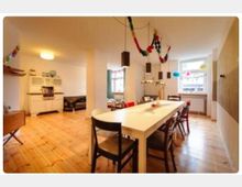 Ein großzügiger Wohn- und Essbereich mit Holzboden, einem langen Esstisch und gemischten Stühlen. Im Hintergrund befinden sich ein Fenster, eine Küchenzeile im Retro-Stil und bunte Dekorationen, darunter Girlanden und Lampions.