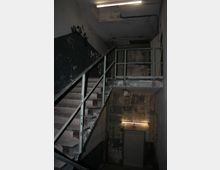 Eine verlassene und heruntergekommene Treppe in einem Gebäude mit abblätternder Farbe, freiliegenden Rohren und bröckelnden Wänden. Die Beleuchtung erfolgt durch Neonröhren, die den Raum spärlich erhellen.