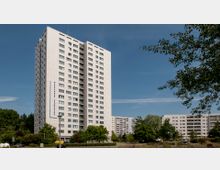 Ein modernes Hochhaus mit weißer Fassade und zahlreichen Fenstern, beschriftet mit „Marzahner Promenade“, umgeben von Bäumen, parkenden Autos und weiteren Wohngebäuden im Hintergrund. Der Himmel ist klar und blau.