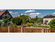 Ein ländlicher Garten mit verschiedenen Blumen, Sträuchern und Gemüsebeeten, umgeben von einem Holzzaun. Im Hintergrund stehen zwei Wohnhäuser mit Giebeldächern und weißen Fassaden, umgeben von Bäumen und Hecken unter einem blauen Himmel mit vereinzelten Wolken.