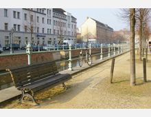 Städtische Szenerie mit einem Kanal, der von einer gepflasterten Promenade mit hellgrünen Geländern gesäumt ist. Im Vordergrund steht eine Holzbank, daneben parkt ein Fahrrad am Geländer, im Hintergrund befinden sich mehrstöckige Gebäude und parkende Autos entlang der Straße.