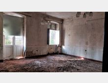 Ein leerer, verlassener Raum mit abblätternden Wänden, einem beschädigten Holzboden und einem offenen Fenster, das Tageslicht hereinlässt. Die Fenster und Türen haben weiße Holzrahmen, während die Decke mit einer alten Lampe versehen ist.