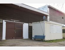 Ein einfacher Gebäudekomplex mit brauner Holzfassade und einer großen, braunen Doppeltür aus Metall, daneben ein beiger Container mit geschlossenen Rollläden und einer blauen Bank davor. Der Untergrund besteht aus sandigem Boden, und im Hintergrund sind Graffiti sowie vereinzelte Äste sichtbar.