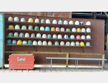 Regal mit mehreren Reihen farbiger Schutzhelme vor einer braunen Metallwand, darunter eine rote Sandkiste mit der Aufschrift „Sand“ und eine schlichte Sitzbank aus Metall. Der Boden besteht aus Pflastersteinen.