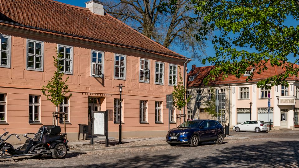 Street view of pastel-colored two-story buildings with red tile roofs and white-framed windows along a cobblestone road, with parked vehicles in front. Leafy trees line the sidewalk, casting shadows across the street.