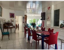 Esszimmer in einem hellen, modern eingerichteten Haus mit einem zentralen Esstisch, umgeben von roten Stühlen. Der Raum zeigt weiße Wand- und Schrankflächen, bunte Wanddekorationen und einen offenen Durchgang zu weiteren Bereichen mit großen Fenstern, die viel natürliches Licht einlassen.