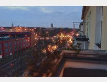 Blick von einem Balkon auf eine beleuchtete Stadtstraße bei Dämmerung, mit einem roten Gebäude und einem REWE-Supermarkt im linken Bildbereich sowie Bahngleisen im Vordergrund. Im Hintergrund sind Wohnhäuser und ein hoher Wasserturm zu sehen.