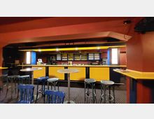 Barbereich in einem Innenraum mit gelb-blauer Theke, Regalen voller Gläser und Spirituosen im Hintergrund. Im Vordergrund stehen hohe Stehtische mit Metallhockern auf einem Teppichboden mit Punktmuster.