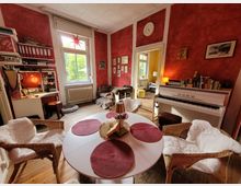 Gemütlicher Wohnraum mit roten Wänden und Holzboden, ausgestattet mit einem runden Esstisch, geflochtenen Stühlen mit Fellauflagen, einem weißen Klavier und einem Schreibtisch vor einem großen Fenster. Im Hintergrund führt eine Tür in einen gelb gestrichenen Raum.