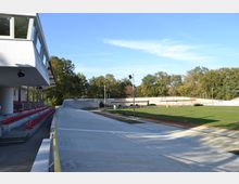 Radrennbahn im Freien mit betonierter Ovalbahn, umgeben von gepflegtem Rasen und Bäumen im Hintergrund. Links im Bild ist eine Tribüne mit roten Sitzreihen und überdachtem Bereich zu sehen.