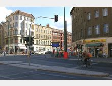 Straßenecke in einem Stadtgebiet mit mehrstöckigen Wohn- und Geschäftsgebäuden, darunter ein Geschäft für Lotto und Tabakwaren. Verkehrsampeln, Fahrräder und eine U-Bahn-Station mit dem Schild „Rosenthaler Platz“ sind sichtbar, während ein Fahrradfahrer die Straße überquert.