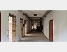 Ein leerer, schmaler Flur in einem scheinbar renovierungsbedürftigen Innenraum mit teilweise offenen Ziegelwänden und unverputzten Türrahmen auf der linken Seite. Im Hintergrund befindet sich eine Treppe aus Metall sowie eine geschlossene Tür mit natürlichem Lichteinfall.