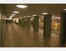 Ein unterirdischer Durchgang mit gepunkteten grünen Stahlstützen, einer beleuchteten Decke und Fliesenboden. Rechts befindet sich eine Glasvitrine mit Zeitschriften und Waren, während Passanten in der Ferne sichtbar sind.