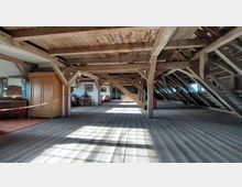 Holzboden-Dachboden mit sichtbaren Holzbalken, spärlich möbliert mit einem Schrank, Spiegel und einigen Sitzgelegenheiten. Tageslicht strömt durch Fenster auf beiden Seiten, wobei ein Seilbereich in der Mitte abgesperrt ist.