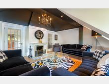 Wohnzimmer in einem Dachgeschoss mit modern-klassischem Interieur, schwarzen Sofas, gemusterten Kissen und einem Glas-Couchtisch auf einem auffälligen Teppich. Im Hintergrund ein Kamin aus weißem Marmor und eine Tür mit Glaseinsatz, die in einen weiteren Raum führt.