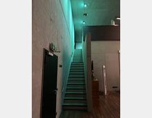 Beton-Innenraum mit schmaler Treppe, die nach oben führt. Links befindet sich eine dunkle Tür mit einem Notausgangsschild darüber, rechts ist ein Bereich mit Holzverkleidung und minimaler Beleuchtung.