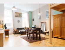 Helle Wohnung mit offener Raumaufteilung, eingerichtet im rustikalen Stil mit Holzmöbeln, einer Sitzgruppe aus Sofa und Sesseln, einem Esstisch mit Stühlen und einem großen Holzschrank. Die Wände sind zum Teil aus Sichtmauerwerk, und zwei große Fenster lassen viel Tageslicht herein.