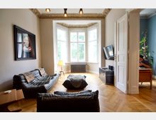 Wohnzimmer in einem Altbau mit hohen Decken und dekorativen Stuckverzierungen. Der Raum verfügt über schwarze Ledersofas, ein großes Fenster mit Fensterbank, ein Bild an der Wand, Holzboden in Fischgrätmuster und einen angrenzenden Arbeitsbereich, der durch eine geöffnete Tür sichtbar ist.