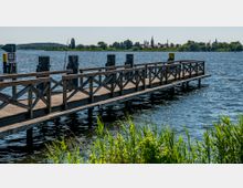 Ein hölzerner Steg ragt in ein großes Gewässer hinein, umgeben von grünem Schilf im Vordergrund. Im Hintergrund sind auf der anderen Seite des Wassers eine kleine Stadt mit Kirchtürmen und Bäumen zu sehen.