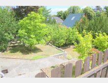 Ein kleiner Garten mit einem gepflasterten Weg, umgeben von grünen Sträuchern und Obstbäumen. Im Hintergrund sind Dächer von Einfamilienhäusern und eine dichte Baumreihe zu sehen, während ein hölzerner Gartenzaun den Vordergrund begrenzt.