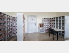 Räumlicher Aktenlagerraum mit mehreren weißen Regalwänden, die mit gestapelten Ordnern und Dokumenten gefüllt sind. Im Vordergrund steht ein Tisch mit einem schwarzen Stuhl, im Hintergrund ist eine weiße Tür sichtbar.