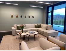Moderne Lounge mit einer großen beige-farbenen Eckcouch, kleinen runden Couchtischen und einer Vase mit Blumen darauf. Im Hintergrund eine pastellgrüne Wand mit kleinen gerahmten Porträtbildern sowie bodentiefe Fenster mit Blick auf eine Außenterrasse und eine winterliche Landschaft.