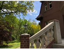 Ein Treppenaufgang mit gemauertem Geländer und Laterne führt zu einem älteren Haus mit rotbrauner Fassade. Im Hintergrund sind ein Garten mit Bäumen und blauer Himmel zu sehen.