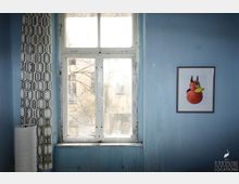 Ein blauer Raum mit einem alten, weißen Holzfenster, durch das Äste und ein benachbartes Gebäude sichtbar sind. Links hängt ein Vorhang mit geometrischem Muster, daneben eine weiße Stehlampe, und rechts an der Wand ein gerahmtes Bild einer roten Gummiente mit Hörnern.