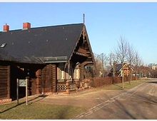 Holzhaus mit dunklem Satteldach und dekorativen Verzierungen an der Fassade, umgeben von einer gepflegten Grünfläche. Im Hintergrund eine ruhige Straße mit weiteren ähnlichen Häusern und kahlen Bäumen.