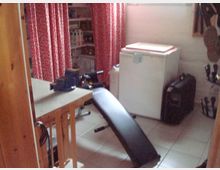 Ein kleiner Raum mit weißen Wänden und Fliesenboden, ausgestattet mit einem Kühlschrank, einer schwarzen Trainingsbank, roten Vorhängen vor Regalen mit gelagerten Gegenständen und einer Werkbank mit Schraubstock.