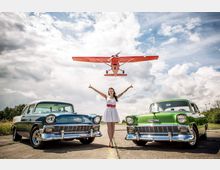 Eine Frau in einem weißen Kleid steht auf einer Straße zwischen zwei klassisch restaurierten Oldtimern, einem blauen und einem grünen Modell, während ein rotes Kleinflugzeug knapp über ihr fliegt. Der Himmel ist teils bewölkt, und die Szene wirkt wie auf einem Flugplatz.