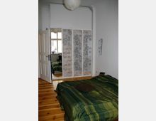 Schlafzimmer mit Holzboden, einem grünen Bettüberwurf und einer hölzernen Tür, die teilweise geöffnet ist. Vor einem großen Spiegel befinden sich weiße Schranktüren mit floralem Muster; eine Papierlampe hängt von der Decke.