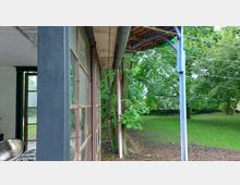 Außenansicht mit einer verglasten Fassade und einem grauen Metallrahmen, daneben ein Überdach mit sichtbarer Metallstütze. Im Hintergrund ist eine Grünfläche mit Bäumen und Sträuchern zu sehen.