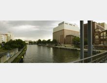 Stadtansicht mit einem Fluss, rechts große backsteinrote und moderne Gebäude, links Bäume und Wohnhäuser. Im Vordergrund führt ein Geländer entlang des Flussufers, das von einer Parkbucht gesäumt ist.