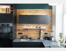 Moderne Küche mit schwarzer und holzgetäfelter Einrichtung. Eine Holzrückwand mit Regal für Gläser und Tassen, eine Arbeitsplatte mit Küchengeräten wie Mixer, Toaster und Wassersprudler, und ein integrierter Backofen in schwarzen Schränken sind sichtbar.