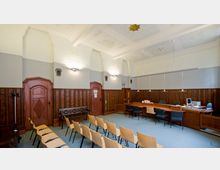 Ein historischer Sitzungssaal mit holzvertäfelten Wänden, hohen Decken mit Stuckverzierungen und hölzernen Türen. Der Raum verfügt über eine Reihe von Holzstühlen, einen langen Tisch mit Bürostühlen sowie Pendelleuchten.