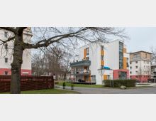 Mehrfamilienhaus in moderner Bauweise mit weißer Fassade und orangefarbenen Akzenten, großen Fenstern und Balkonen. Im Vordergrund ein Baum mit kahlen Ästen sowie ein Weg und etwas Grünfläche; im Hintergrund weitere Wohngebäude.