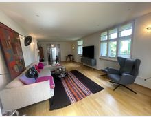 Helles Wohnzimmer mit Holzfußboden, großem Sofa mit Couchtisch und farbigem Teppich sowie einem Fernseher auf einem Lowboard. Rechts sorgen hohe, mehrteilige Fenster für viel Tageslicht; daneben steht ein gepolsterter Sessel, im Hintergrund führt eine Glastür in den nächsten Raum.
