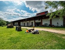 Ländliches Hofgelände mit einem langen, niedrigen Wirtschaftsgebäude mit weißer Fassade und dunklem Dach, davor ein Schotterweg und Grünfläche. Im Vordergrund liegt ein kleiner Haufen Material auf dem Rasen; im Hintergrund sind Bäume und ein wolkiger Himmel zu sehen.