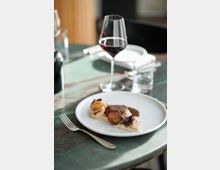 Ein Teller mit einem raffinierten Gericht, bestehend aus gebratenem Fleisch, Kartoffelscheiben, Soße und Dekoration aus essbaren Kräutern, steht auf einem grünen Marmortisch. Daneben befinden sich ein Weinglas mit Rotwein, eine Gabel und ein Wasserglas aufgedeckt.