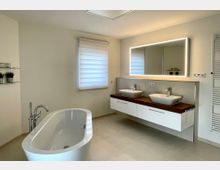 Modernes Badezimmer mit freistehender Badewanne und großem Doppelwaschtisch auf Holzplatte. An der Wand hängt ein breiter, beleuchteter Spiegel über weißen Unterschränken; daneben sind ein Fenster mit Rollo und ein Handtuchheizkörper zu sehen.