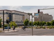 Straßenkreuzung mit Ampeln und Straßenschildern, die "Allee der Kosmonauten" und "Marzahner Chaussee" anzeigen, im Vordergrund. Im Hintergrund befinden sich zwei große, rechteckige Plattenbauten, umgeben von Bäumen und parkenden Autos.