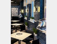 Modern eingerichtetes Café mit Tischen aus weißem Marmorimitat, schwarzen Stühlen und einer Wandverkleidung aus glänzenden dunkelblauen Fliesen. An der Wand befinden sich rechteckige Wandleuchten und Fenster mit kleinen Pflanzen in weißen Töpfen auf den Fensterbänken.