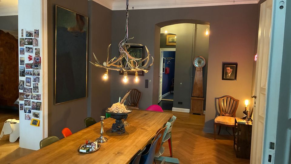 Esszimmer mit langem Holztisch und unterschiedlich farbigen Stühlen, darüber eine hängende Geweih-Lampe mit sichtbaren Glühbirnen. Graue Wände mit großen Bildern, Stuckdecke und Fischgrätparkett; im Hintergrund ein Rundbogen-Durchgang und eine Standuhr.