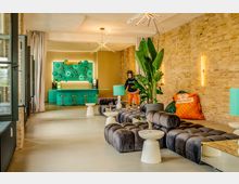 Moderner Lounge- und Barbereich in einem Loft mit Sichtziegelwand, großen schwarzen Polstersofas und kleinen weißen Beistelltischen. Im Hintergrund steht eine türkisfarbene Bar mit Hockern und beleuchtetem Flaschenregal; dekorative Deckenleuchten, hohe Pflanzen und Vorhänge rahmen den Raum.